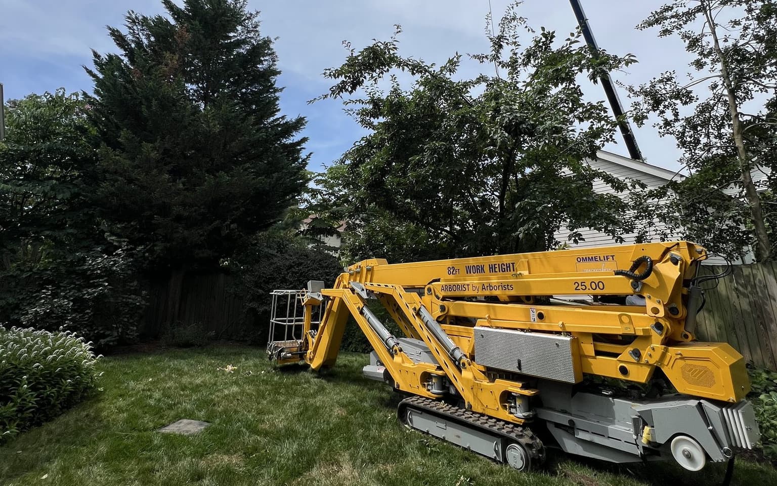 A yellow Ommelift tracked spider lift set up on a lawn with its outriggers down, the crane’s cable visible above it and a wooden fence behind