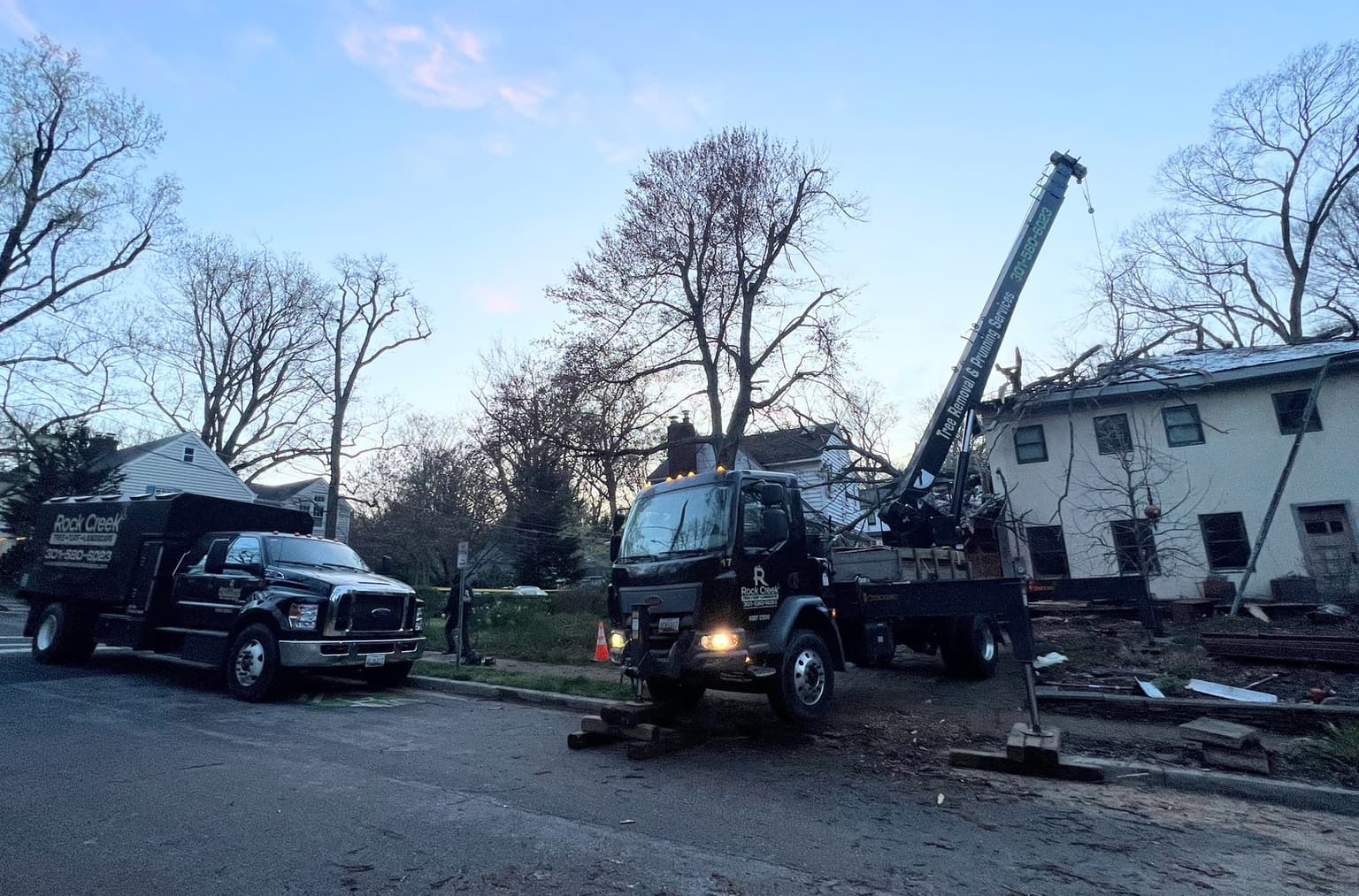 A Rock Creek boom truck and chip truck set up in a residential street at dusk with a traffic cone out, working on a storm-damaged house