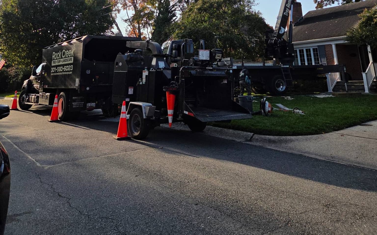 The chipper and chip truck coned off along the street, with the crane’s boom rising from in front of the house to the right